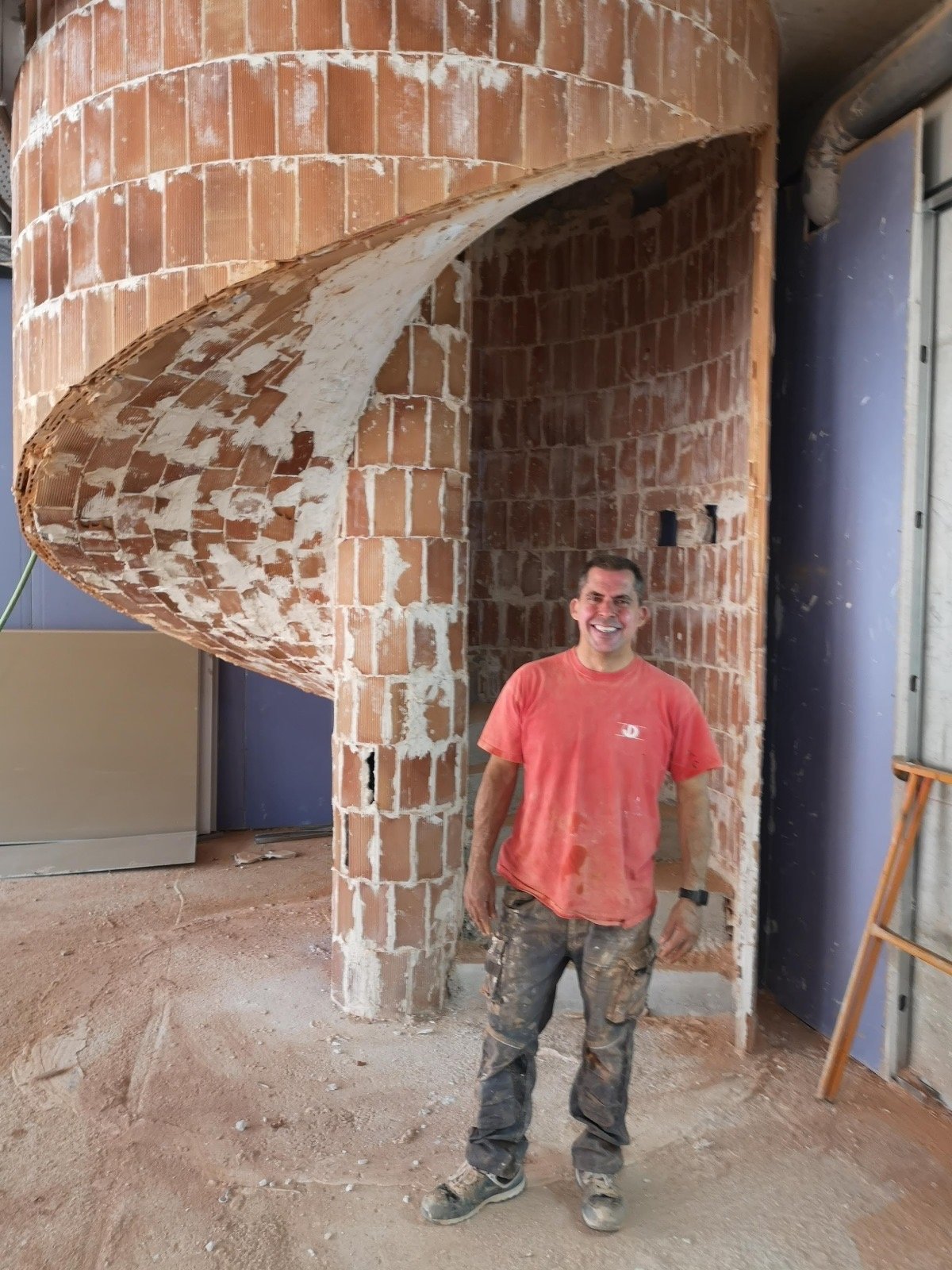 Hombre con camiseta roja y pantalones de trabajo manchados de polvo posando frente a una escalera de caracol construida con ladrillos, dentro de un edificio en construcción.