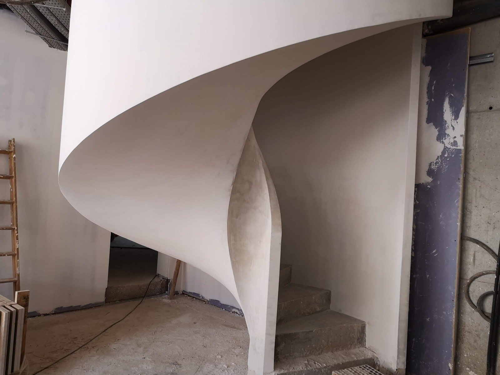Escalera de caracol de hormigón con barandilla curva enlucida de color blanco, vista desde la base, con peldaños de concreto sin acabado y materiales de construcción apoyados en la pared al fondo.