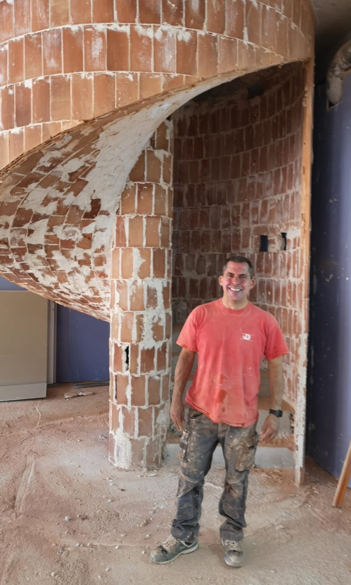89186567-00f9-4390-8aa1-9572e39e35bb Hombre con camiseta roja y pantalones de trabajo manchados de polvo posando frente a una escalera de caracol construida con ladrillos, dentro de un edificio en construcción.