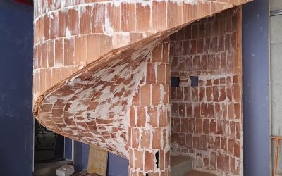 Escalera de caracol de ladrillo sin enlucir, en fase de construcción, con barandilla curva y escalones visibles, rodeada de polvo y materiales de obra.